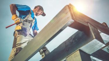 Handwerker steht auf einem Gerüst, hält einen Hammer und trägt einen Gürtel mit Werkzeug in einer Baustellenszene.