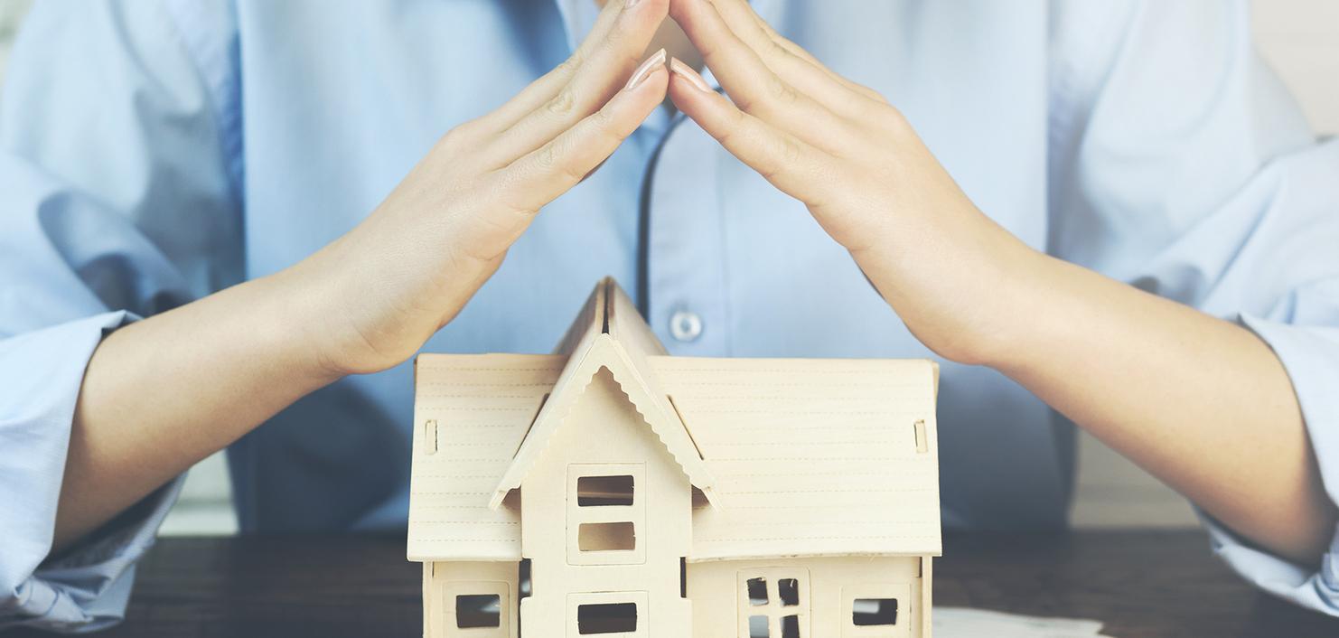 Hände einer Person legen schützend über ein kleines Holzhaus auf einem Tisch, daneben ein Sparschwein und eine Lupe.